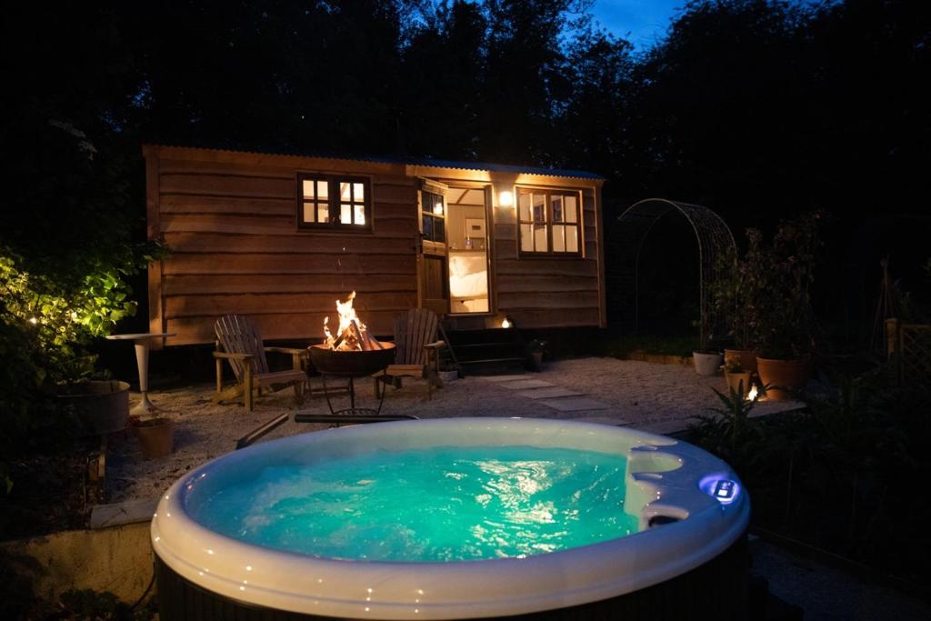 Luxury Shepherds Hut near Bristol. This is night time poto with a wooden converted shepherds hut in the back ground and an illuminated hot tub in the foreground. There is a lit fire pit with 2 chairs